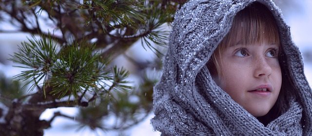 Flicka med stickad sjal framför en tall. I bakgrunden och på tallen syns även snö.